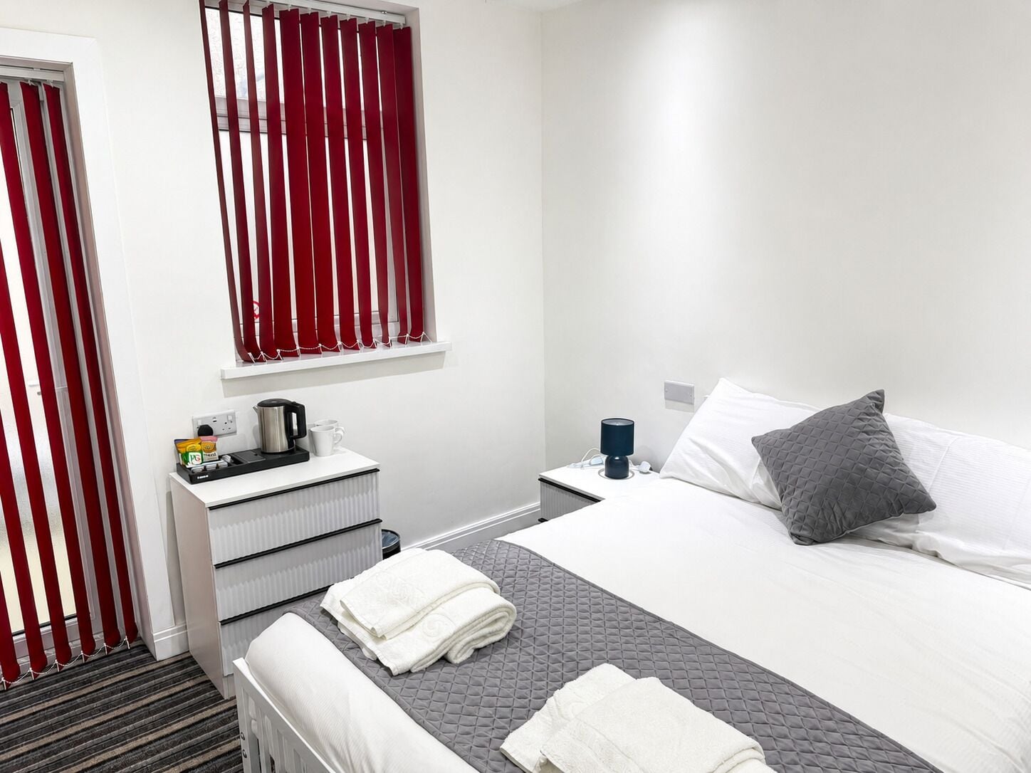 A hotel room with a bed, bedside table, and a window with red curtains.