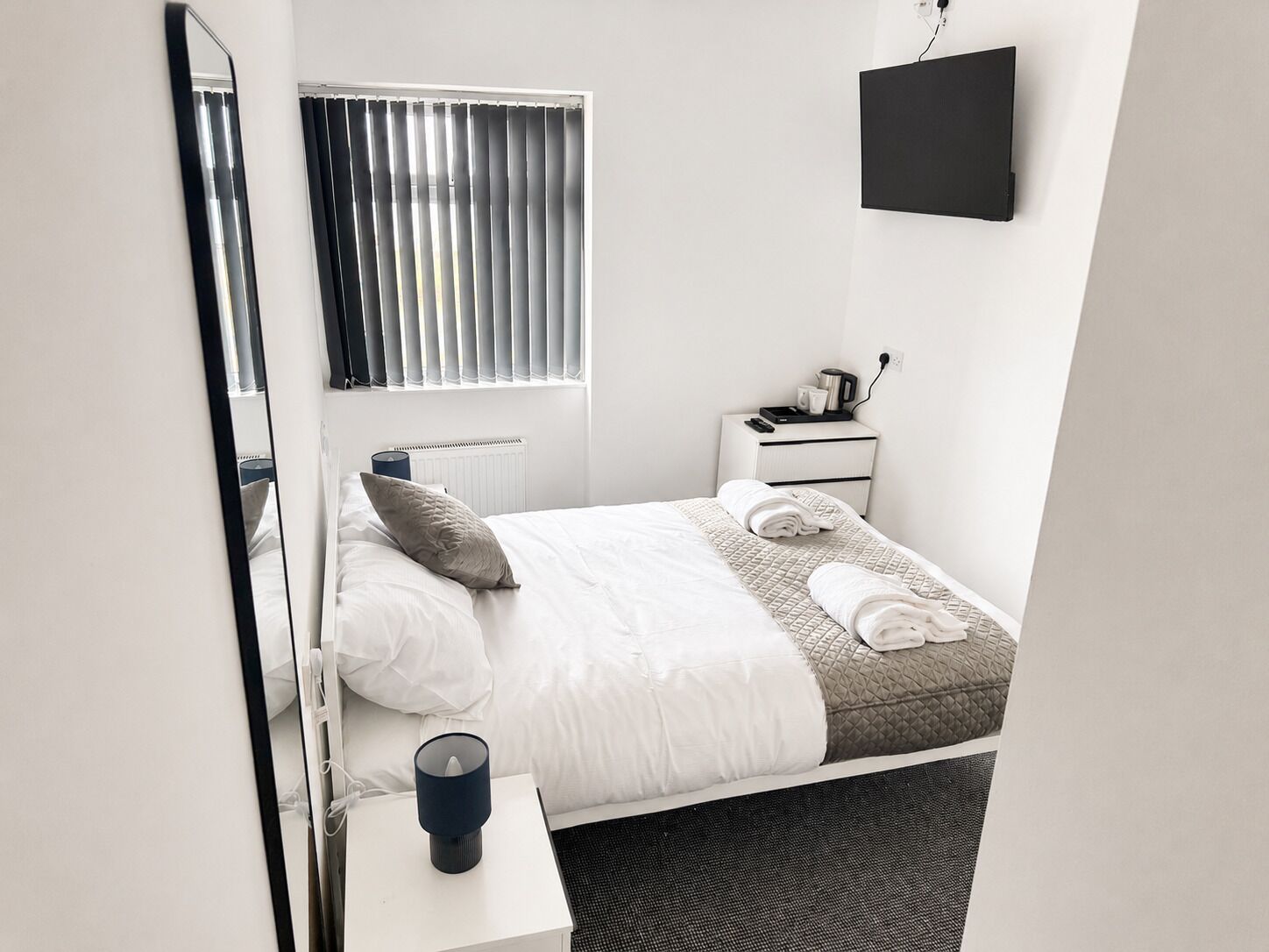 Quarto de hotel com cama, mesa de cabeceira com abajur, espaço para café e televisão fixada na parede, além de uma janela com persianas.