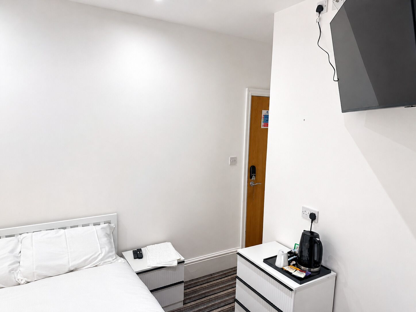 A hotel room with a bed, a bedside table with a remote control, a coffee maker, and a wall-mounted television.