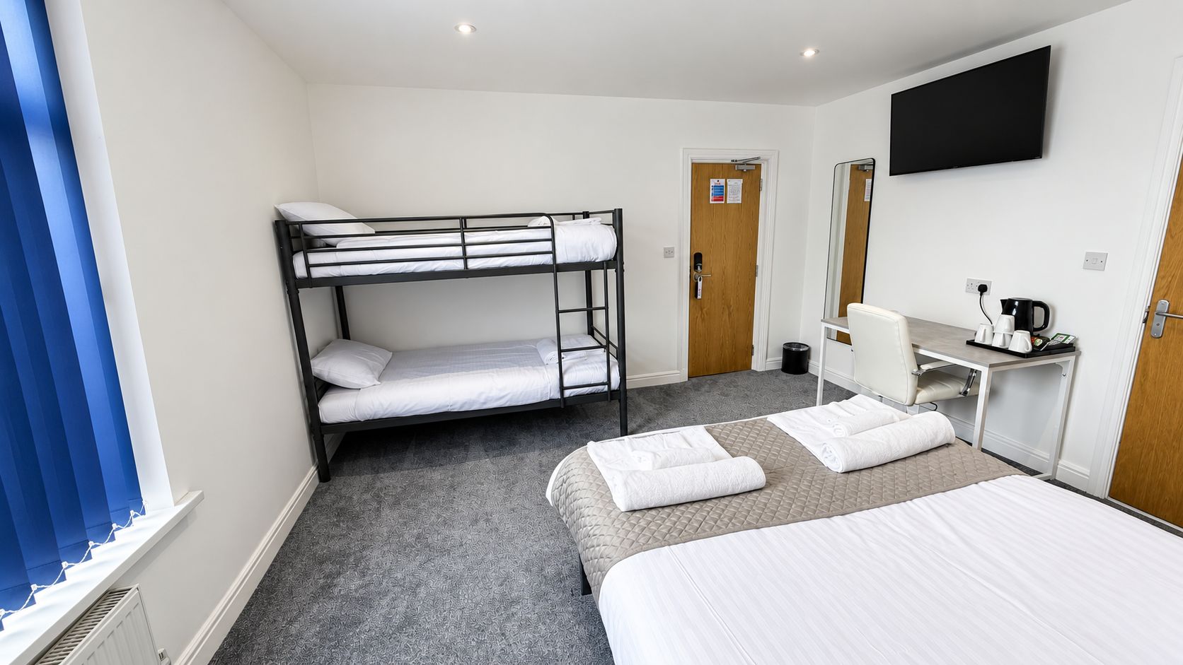 A hotel room with a bunk bed, a single bed, a desk with a chair, a television, and a coffee machine.