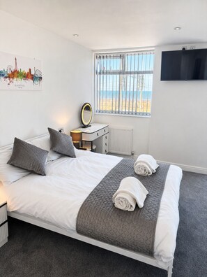 A hotel room with a bed, a bedside table with a mirror, a TV mounted on the wall, and a window with a view of the beach. - 399 Prom - Blackpool Hotel (Blackpool)