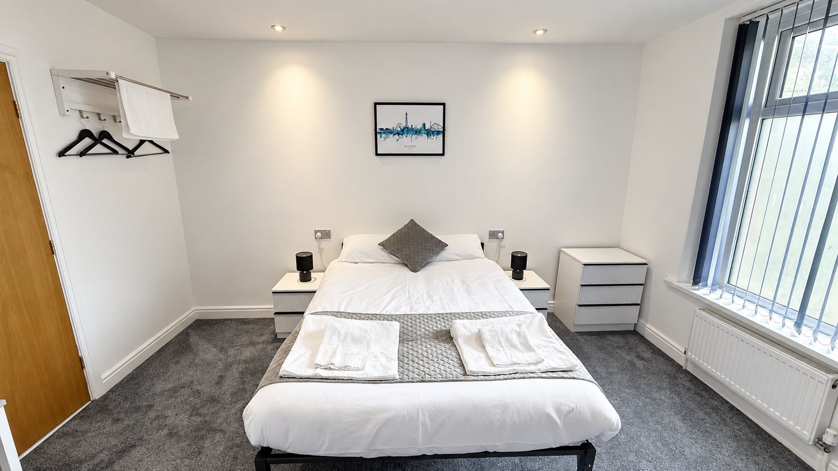 A neatly arranged bedroom with a bed, bedside tables, a window with blinds, a wall-mounted towel rack, and a cityscape artwork.