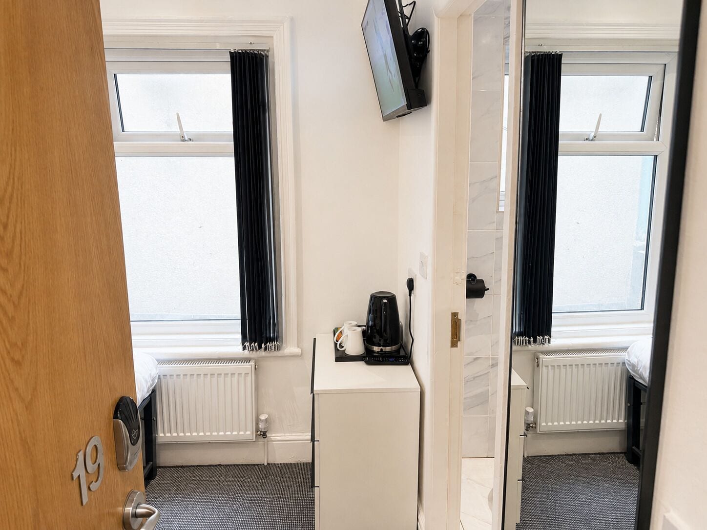 A hotel room with two windows, a white radiator, a small white cabinet, and a television mounted on the wall.