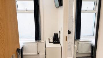 A hotel room with two windows, a white radiator, a small white cabinet, and a television mounted on the wall.