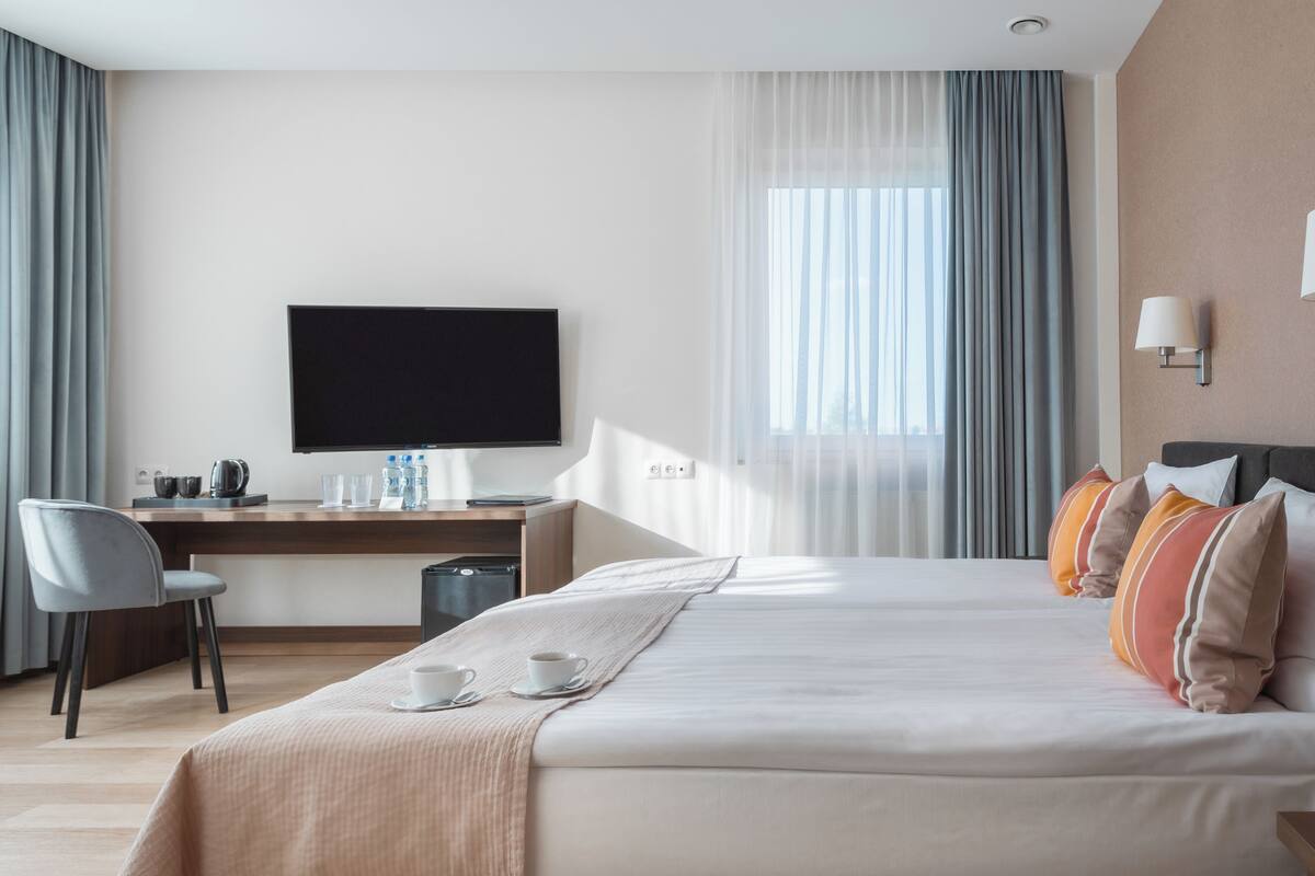 A hotel room with a bed, a desk with a flat-screen TV, a chair, a coffee table with cups, and a window with curtains.