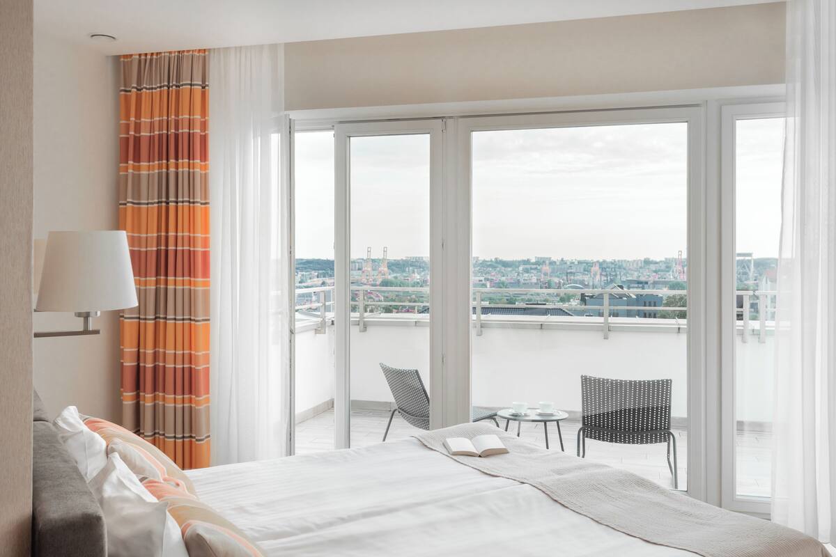 A hotel room with a bed, a chair, a small table, and a view of a cityscape through large windows.