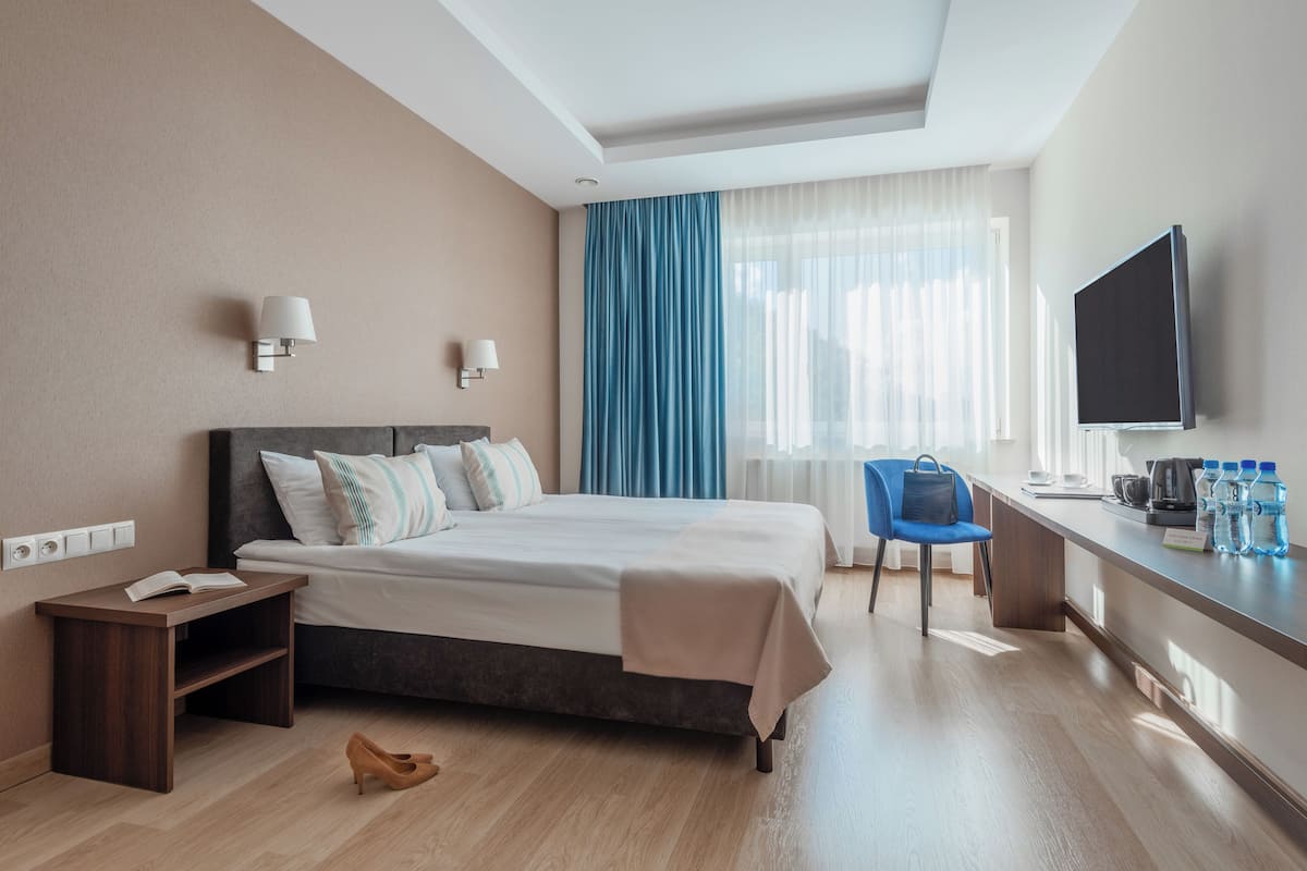 A hotel room with a bed, bedside table, chair, TV, and a desk with water bottles.