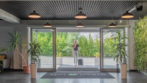 L’entrée d’un hôtel, avec une porte en verre, des plantes en pot et une personne tenant une valise.