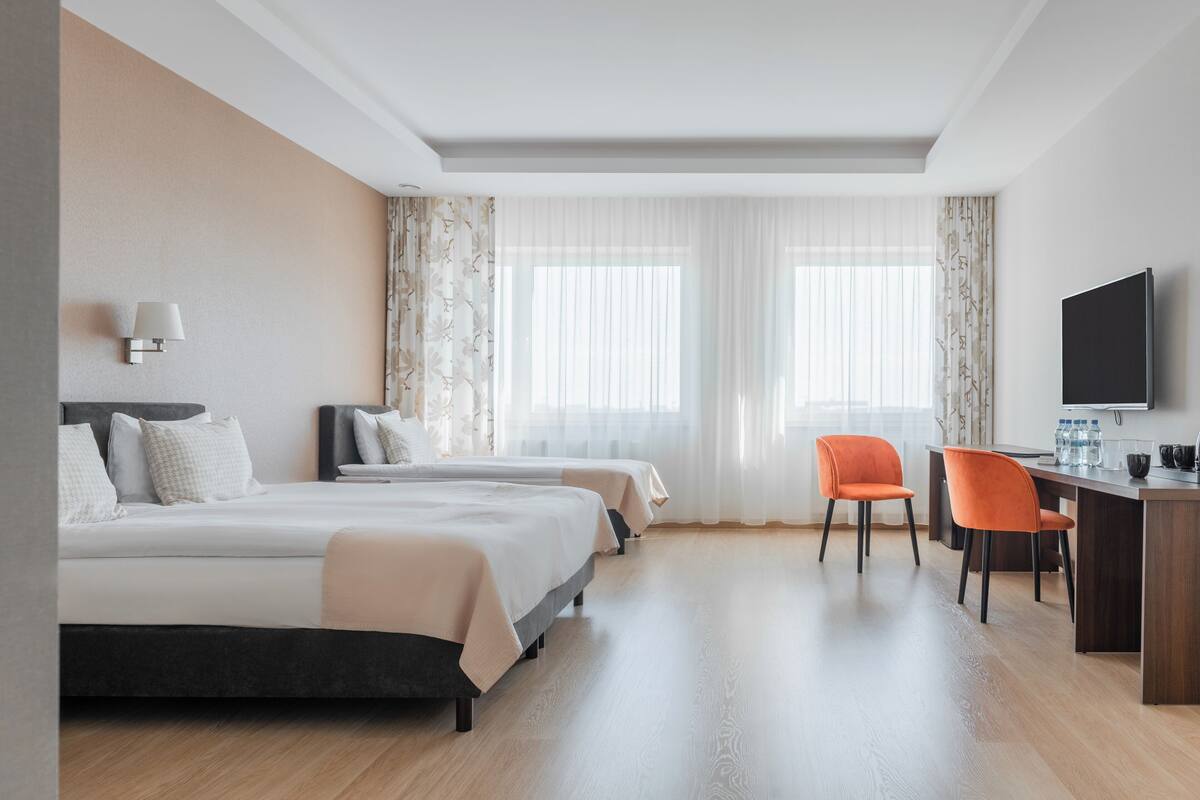 A hotel room with two beds, a desk with chairs, a TV, and large windows with sheer curtains.