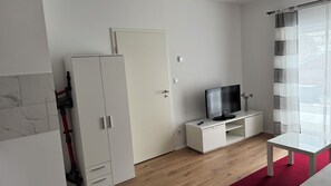 Huoneessa on valkoinen vaatekaappi, valkoisella jalustalla oleva televisio, punainen matto ja ikkunassa kuviollinen verho.