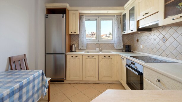 A compact kitchen with a refrigerator, a sink, and a dishwasher. There is a window with curtains, a table with a checkered tablecloth, and a chair. - Apartment 'Casa Verde Blu' with Balcony, Wi-Fi and Air Conditioning (Perivoli)