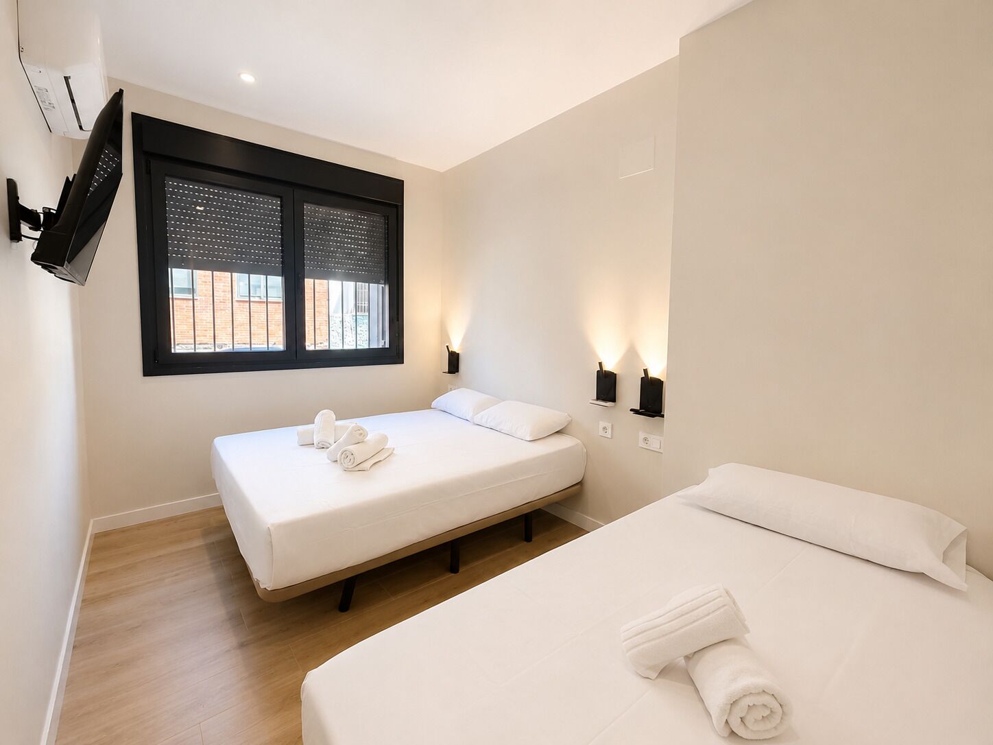 A hotel room with two beds, a TV mounted on the wall, and a window with blinds.