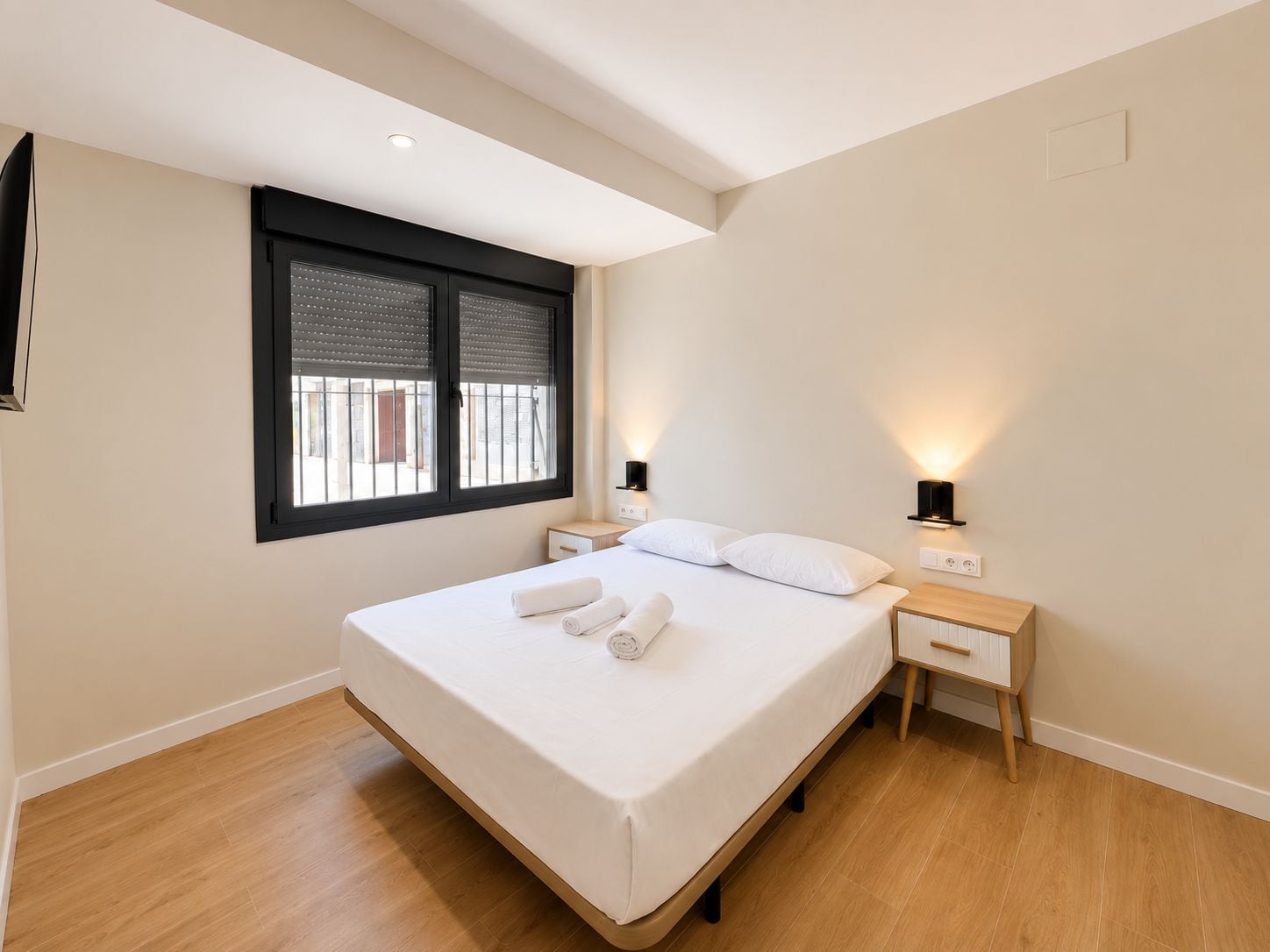 Un dormitorio moderno con una cama grande, mesitas de noche, una ventana con persianas y luces fijadas a la pared.