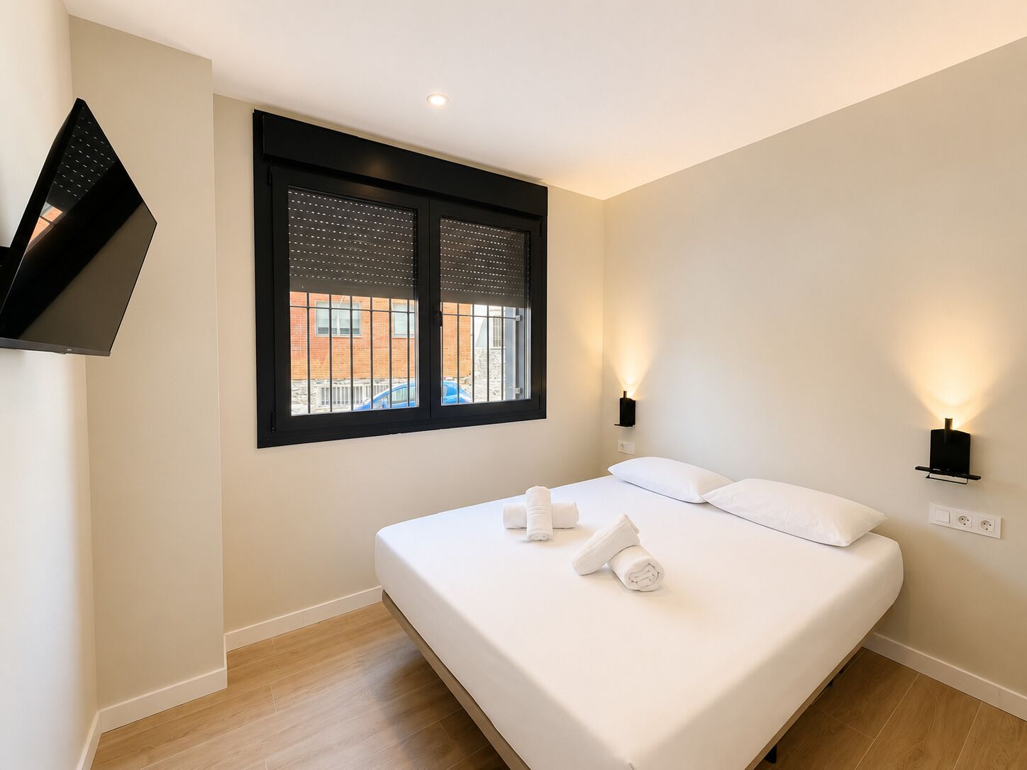 Una habitación de hotel moderna con una cama, dos lámparas de noche, un televisor montado en la pared y un ventanal con persianas.