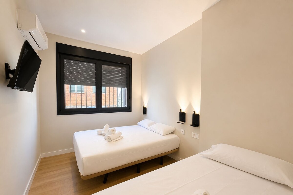 A hotel room with a bed, a TV mounted on the wall, a window with blinds, and a wall-mounted light fixture.