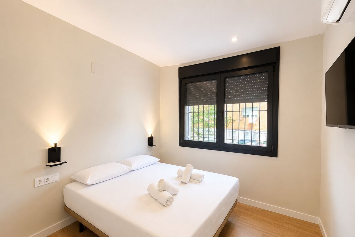A hotel room with a bed, two wall-mounted lights, a window with blinds, and a flat-screen TV.