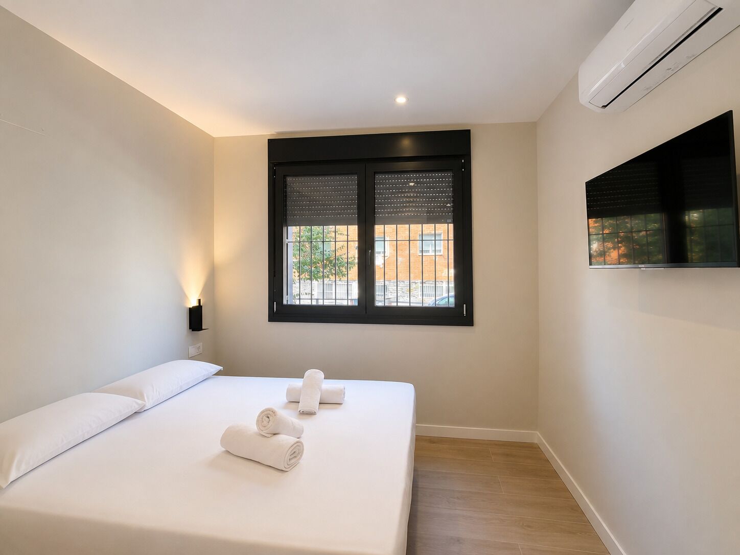 A hotel room with a bed, a window with blinds, an air conditioning unit, and a wall-mounted TV.
