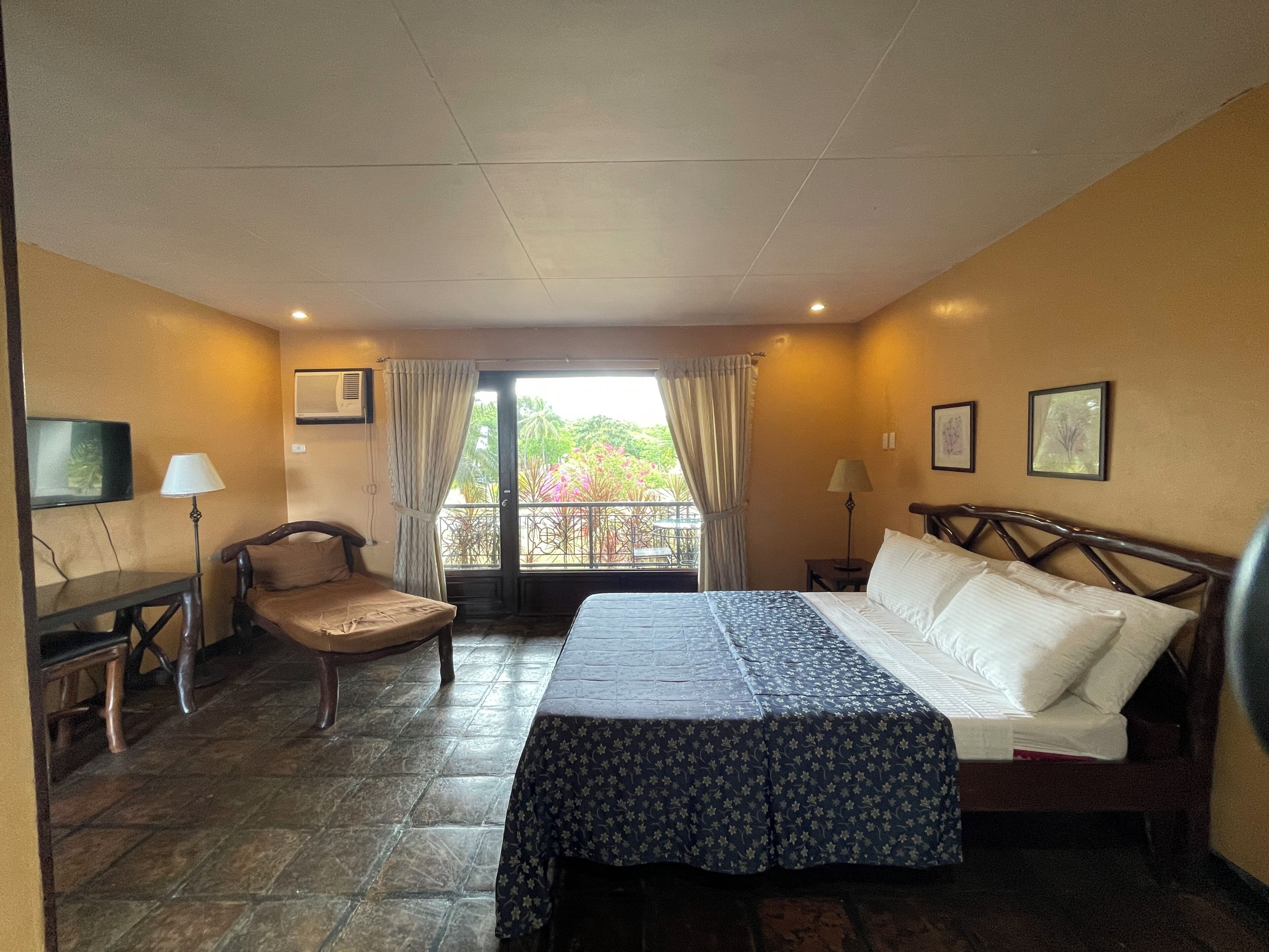 Una habitación de hotel con cama, una silla, un escritorio, televisor y un balcón con cortinas.
