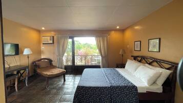 Una habitación de hotel con cama, una silla, un escritorio, televisor y un balcón con cortinas.