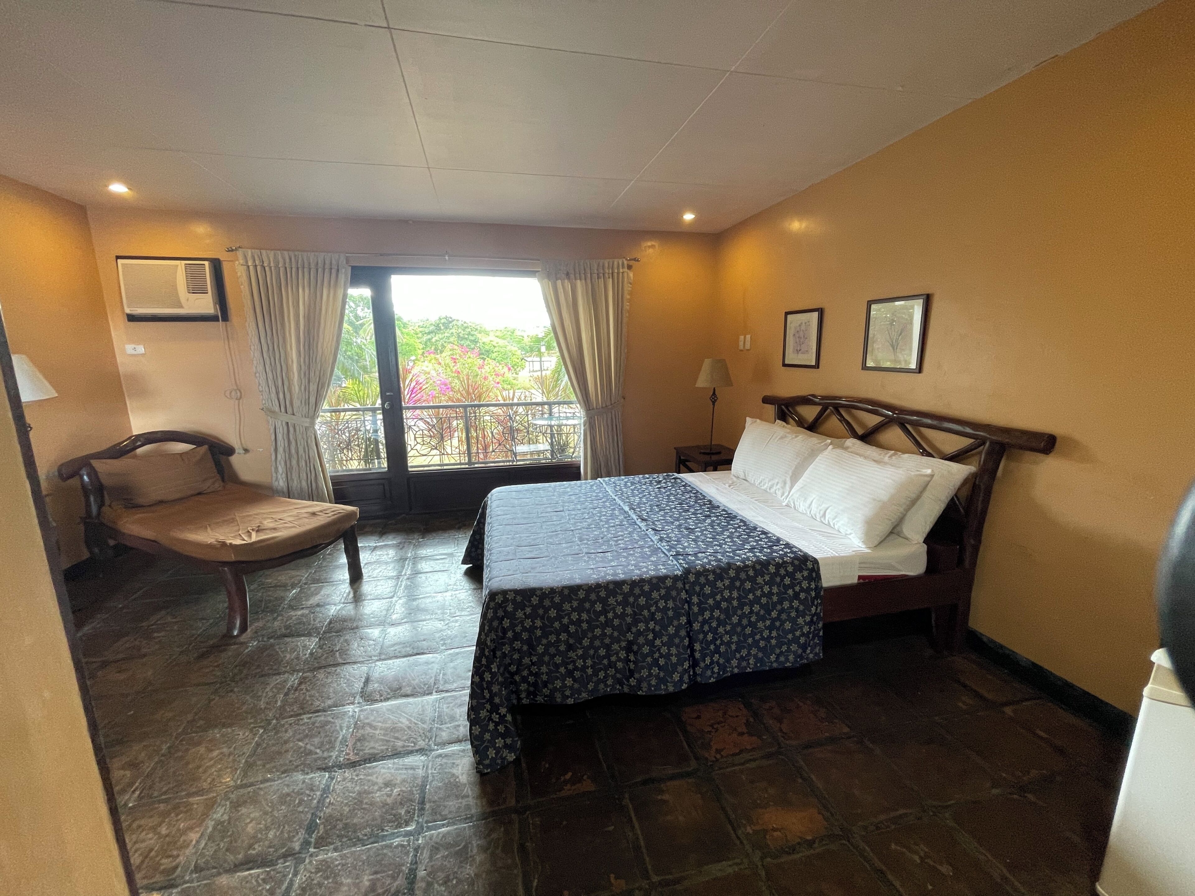 Habitación de hotel con cama, una silla, mesita de noche, ventana con cortinas y vista a zonas verdes.