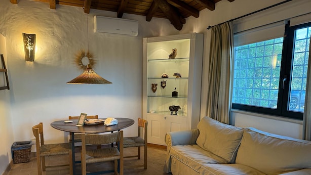 Una habitación acogedora con una mesa redonda de madera, un sofá con cojines, un aparador de vidrio con adornos y una ventana con vista a la vegetación.