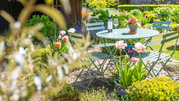 A garden setting with a round table, chairs, and a bottle of wine.