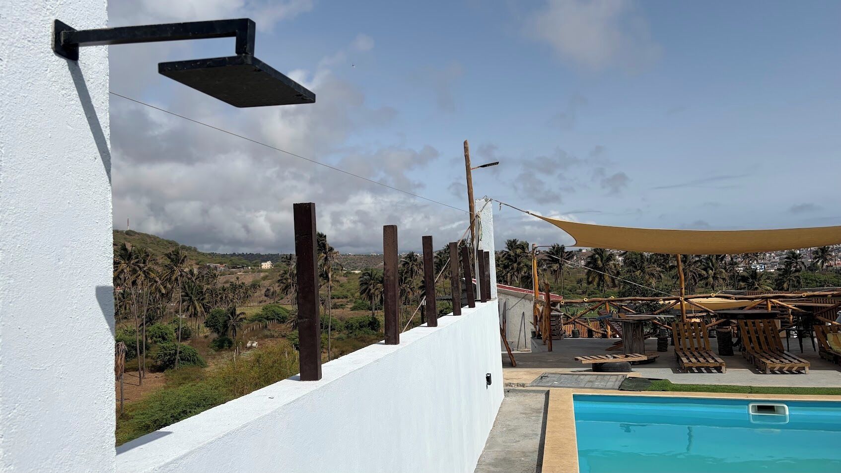 A poolside area with a white wall, wooden deck, and a view of palm trees and hills.