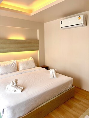 Habitación de hotel con cama, ropa blanca, cabecera de madera, equipo de aire acondicionado y mesita de noche con lámpara.