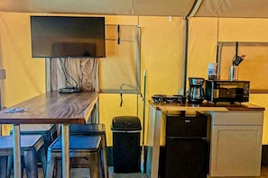 A compact kitchenette with a microwave, coffee maker, and a small refrigerator. There is a table with chairs and a TV mounted on the wall. - Discover Glamping at its Best in this Lovely Safari Tent Rental in the Heart of Texas (Cibolo)