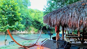 A hammock, a grill, and a thatched-roof structure by a river. - Fantastic Glamping Getaway near Kingsbury, Texas: Beautiful Fully-Equipped Safari Tent on the San Marcos River (Kingsbury)