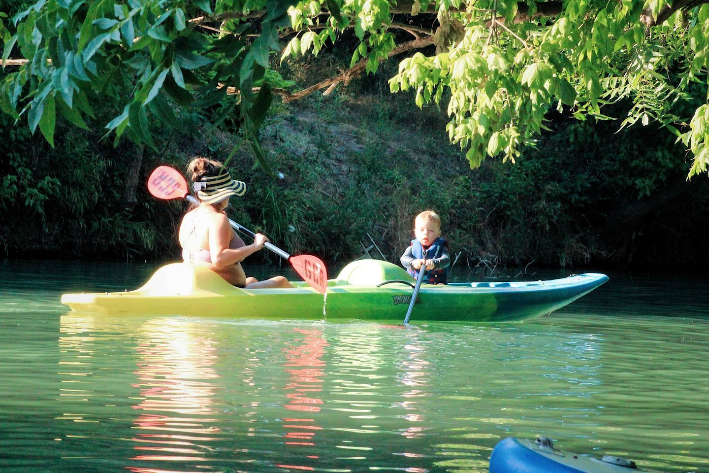 Two people kayaking on a calm river surrounded by greenery.