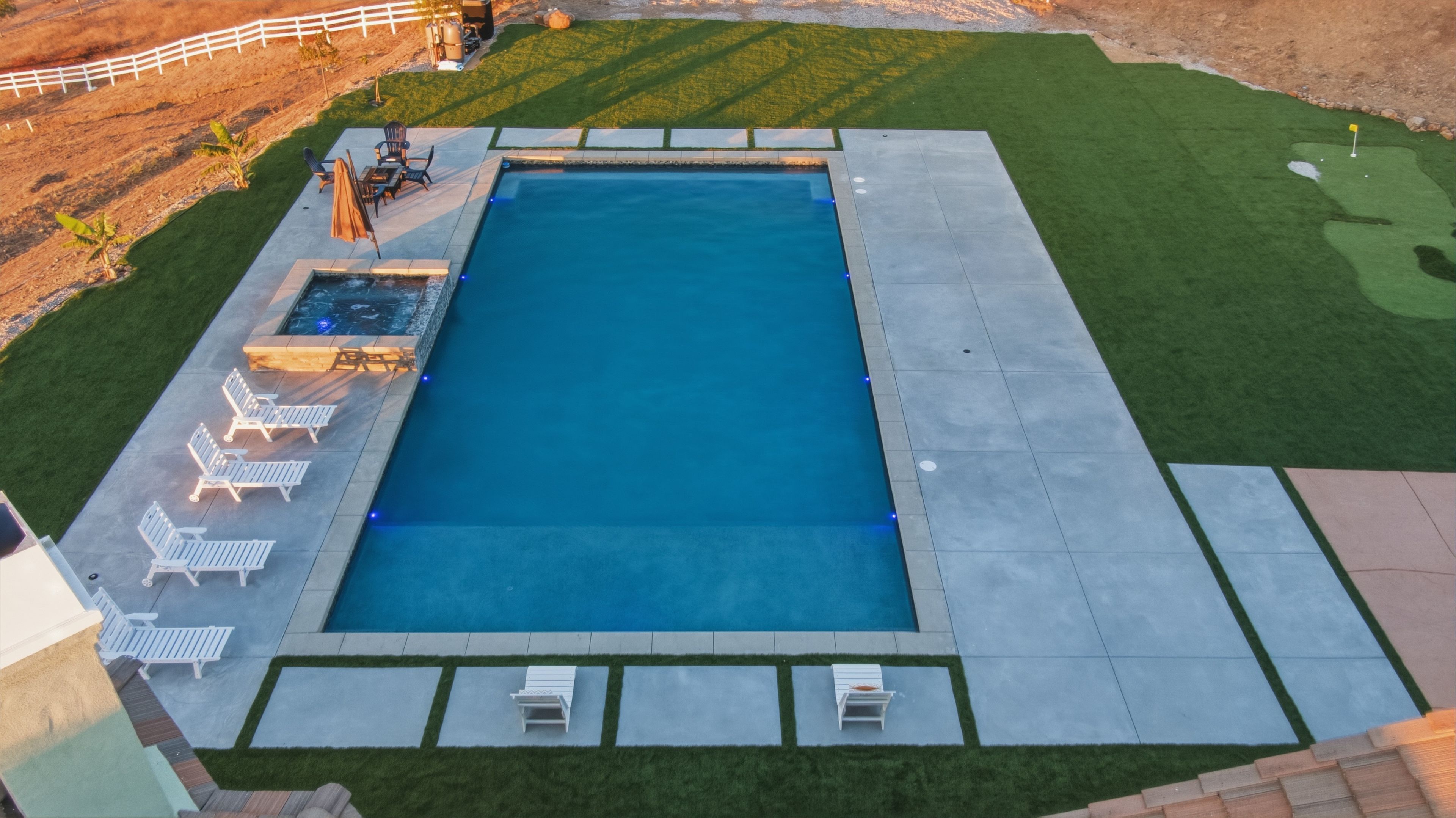 A rectangular swimming pool with a surrounding deck, lounge chairs, and a small table.