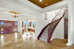 A spacious foyer with a grand staircase, a chandelier, and a view through the open doors.