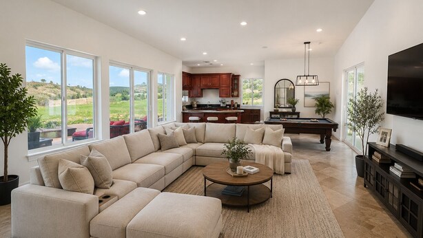 A spacious living room with a large sectional sofa, a coffee table, and a pool table. There is a kitchen area with wooden cabinets and a large window overlooking a scenic landscape.