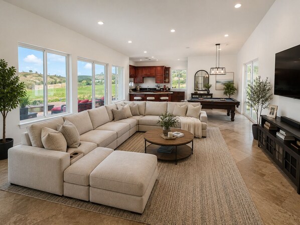 A spacious living room with a large sectional sofa, a coffee table, and a pool table. There is a kitchen area with wooden cabinets and a large window overlooking a scenic landscape.