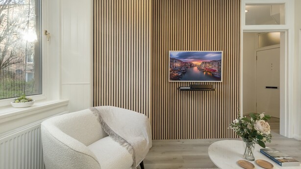 Una sala de estar moderna con un sofá, un televisor de pantalla plana montado en una pared de paneles de madera, una mesita con un florero y una ventana con vista a edificios.