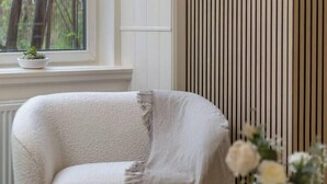 Una habitación con ventana, una silla, una mesa con un florero y un panel vertical.