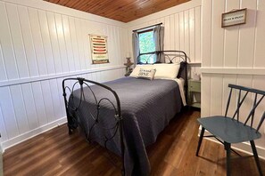 A bedroom with a bed, a chair, a window with curtains, a wall-mounted sign, and a wall decoration. - Charming Waterfront Cabins with Hot Tub & Private Beach Access near Traverse City, Michigan (Traverse City)