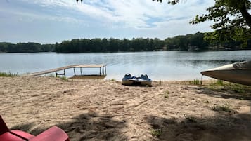 A sandy beach with a wooden dock, two boats, and red chairs.
