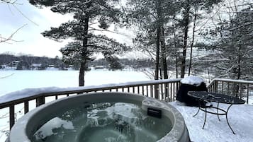 A hot tub on a snow-covered deck with a view of a lake and trees.