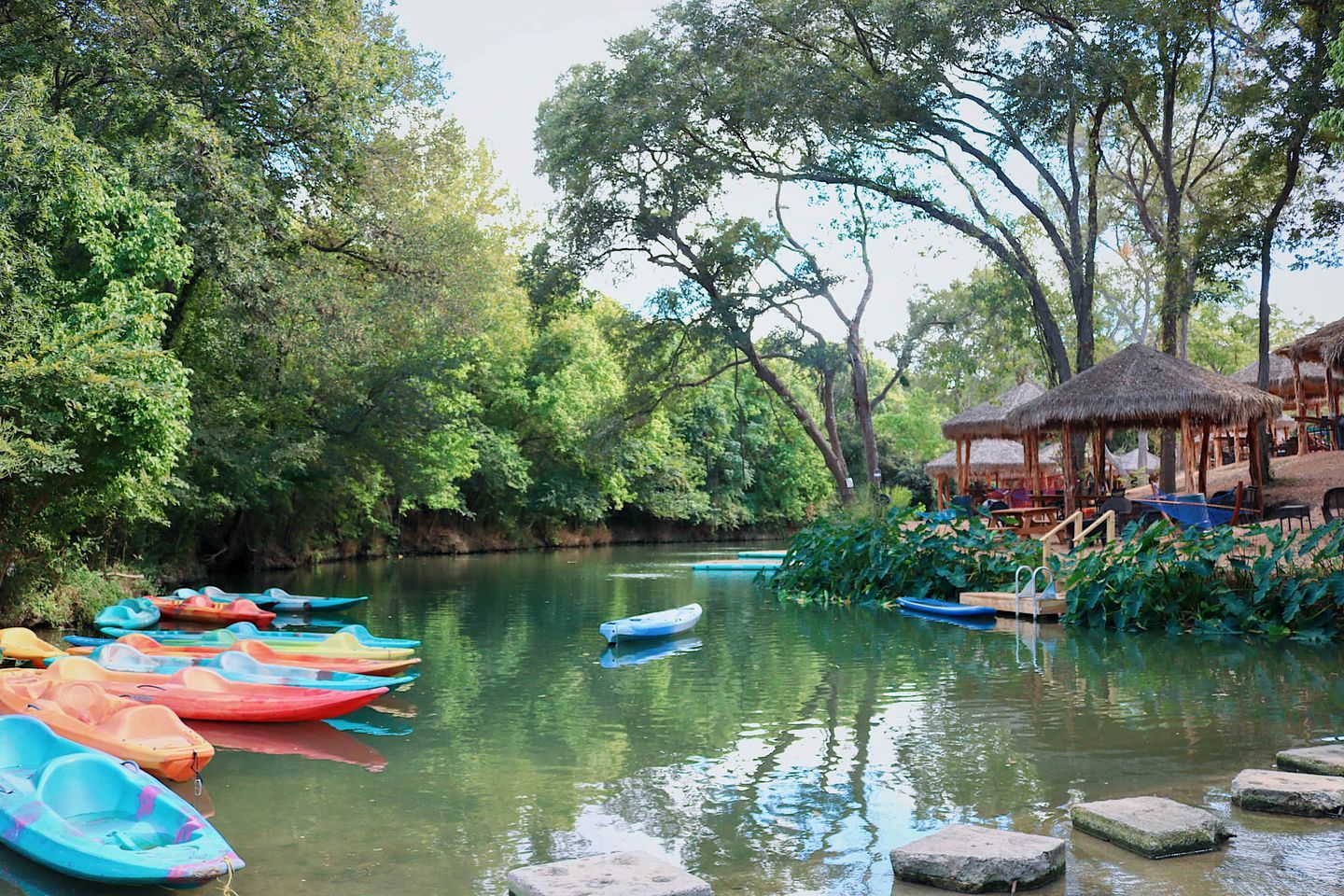 A calm river with colorful boats, a thatched-roof structure, and lush greenery.