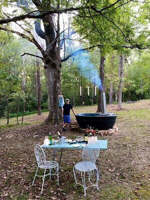 A table with chairs in a forested area, a person standing, and a barbecue grill emitting smoke. - Glamping Tiny House Situated in the Tennesse Woods for a Tranquil Escape near Chattanooga (Signal Mountain)