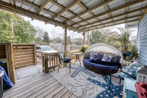 A covered patio with a wooden deck, a wicker sofa with cushions, a small table, and a chair. - Cozy Blue Beach Bungalo • Walk to Shore • (Gulfport)