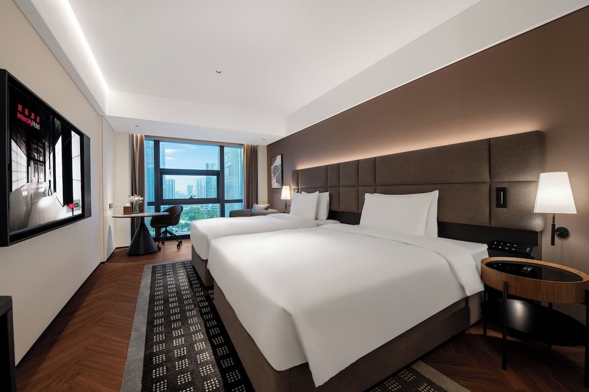 A hotel room with two beds, a desk, a chair, a TV, and a large window with a city view.