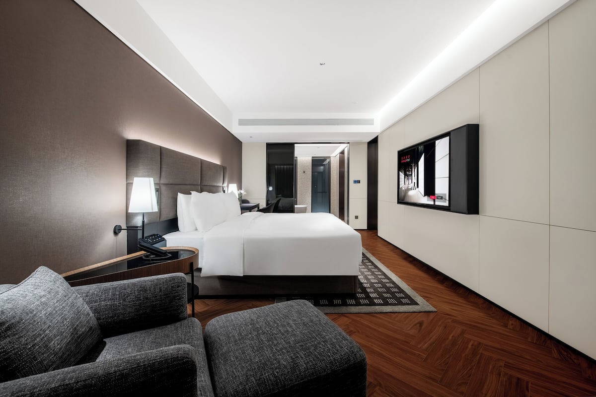 A modern hotel room with a large bed, a desk, a chair, a lamp, and a television.