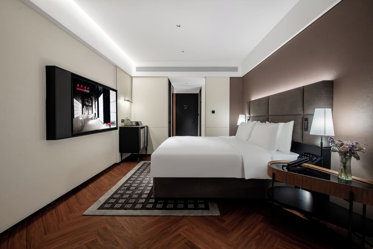 A modern hotel room with a large bed, a bedside table with a lamp, a small desk, and a wall-mounted TV.