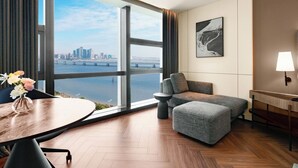 Una habitación de hotel moderna con un ventanal grande, una mesa de comedor con un florero, un sofá gris y una lámpara.