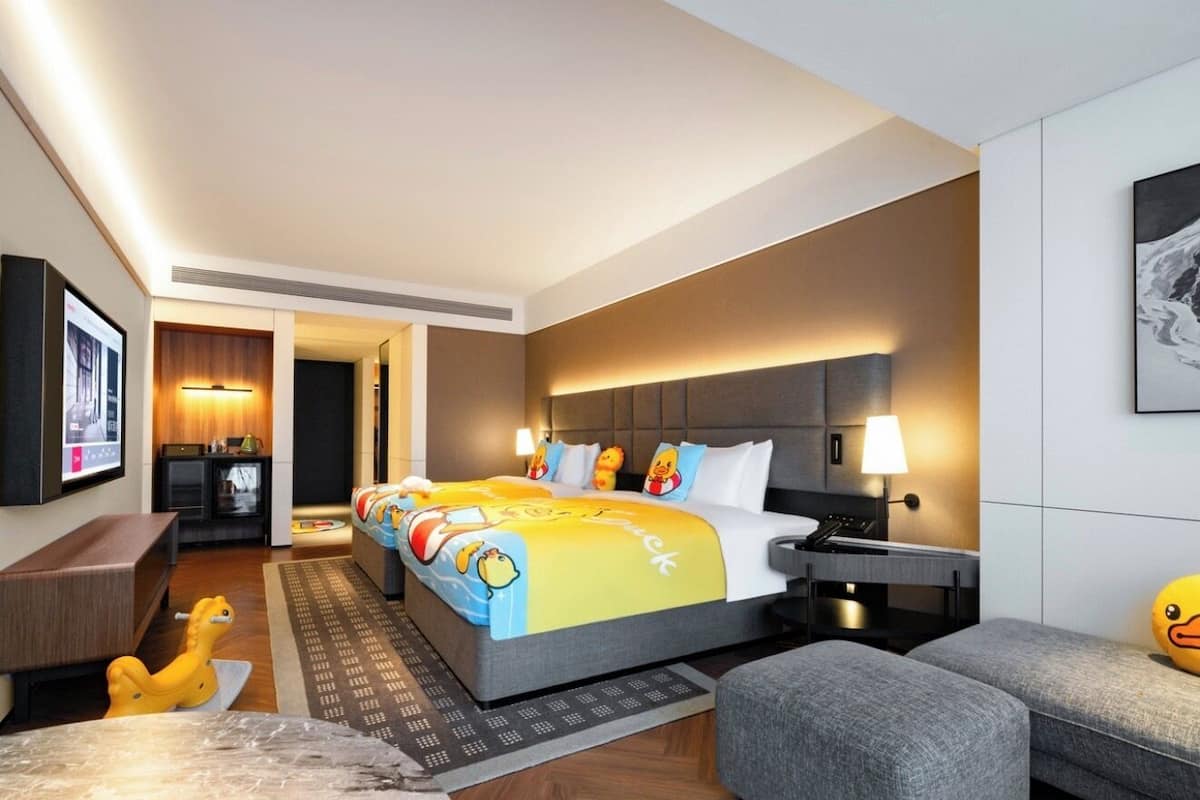 A hotel room with a large bed, a desk, a chair, a sofa, a TV, and a lamp.