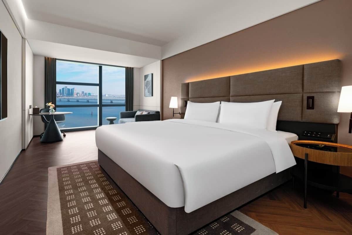 A modern hotel room with a large bed, a desk, a chair, a lamp, and a view of the city through large windows.