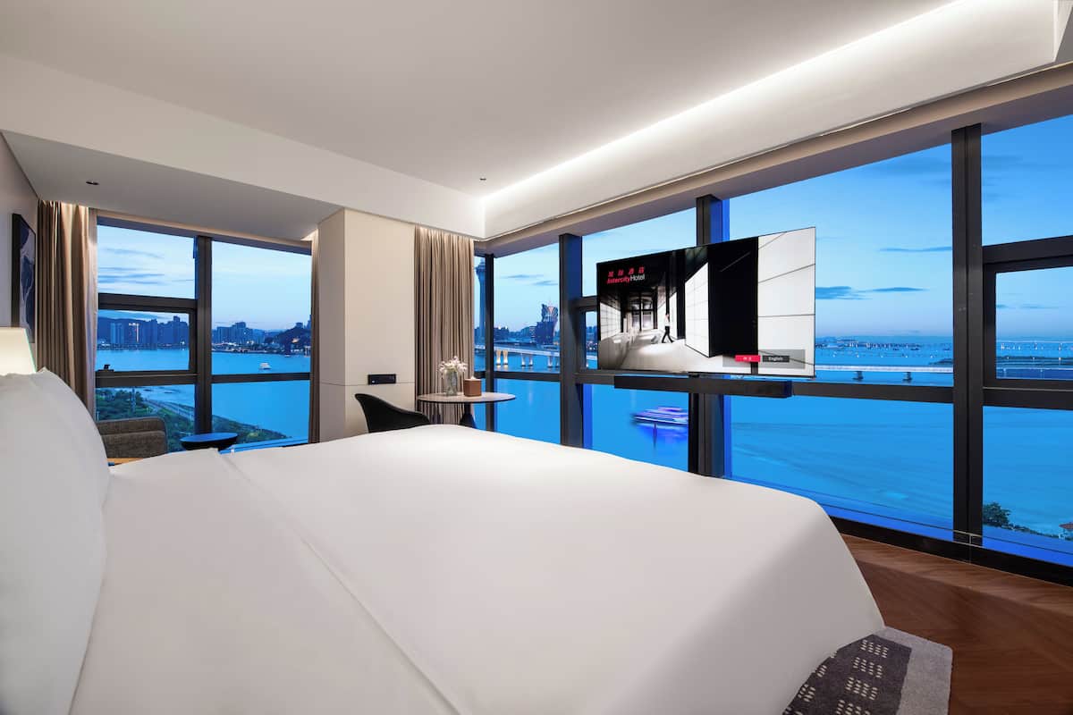 A modern hotel room with a large bed, a TV, a small table, and a view of the city and water.